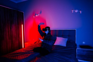 Genç bir kadın gece yatak odasında yatağında otururken sanal gerçeklik gözlüğü kullanıyor.