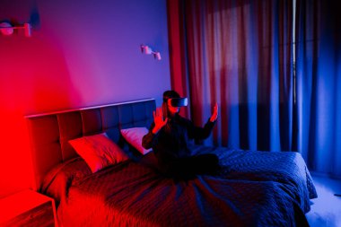 Genç bir kadın loş bir odada yatağında otururken sanal gerçeklik gözlüğü kullanıyor.