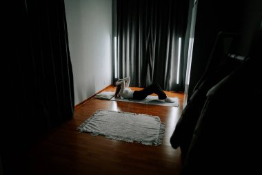 Genç bir kadın, perdeleri ve yerde paspası olan loş bir odada egzersiz yapıyor.