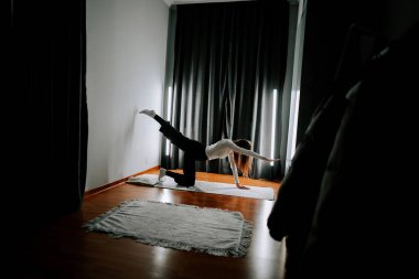 Genç bir kadın, siyah perdeleri ve ahşap zemini olan bir odada paspasın üzerinde egzersiz yapıyor.