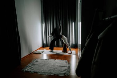 Genç bir kadın akşam saatlerinde oturma odasında perdeleri kapalı olarak egzersiz yapıyor.