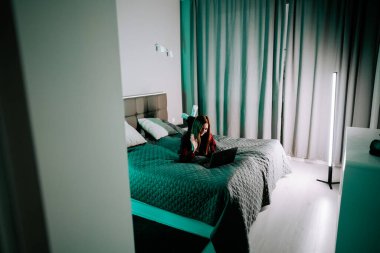 Genç bir kadın akşam saatlerinde rahat bir odada dizüstü bilgisayarla ders çalışıyor.