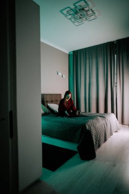 Genç kadın laptopta çalışırken modern yatak odasında hafif ışıklandırmayla oturuyor.