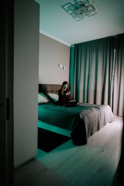 Genç bir kadın akşam saatlerinde rahat bir odada yatağında otururken dizüstü bilgisayarla ders çalışıyor.