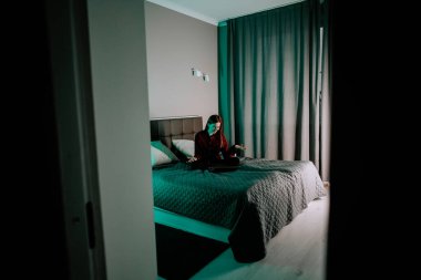 Genç bir kadın akşam saatlerinde odasında yatarken dizüstü bilgisayarda ders çalışıyor.