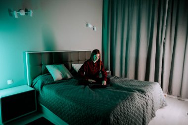 Genç kadın, akşam saatlerinde rahat bir odada yatarken dizüstü bilgisayarda ders çalışıyor.