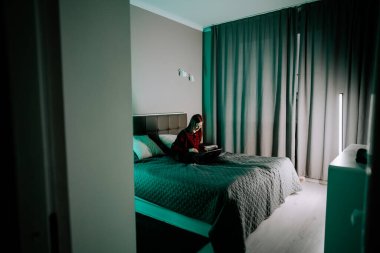 Genç bir kadın, perdeleri kapalı bir odada yatarken dizüstü bilgisayarda ders çalışıyor.