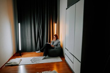 Genç bir kadın, perdeleri ve ışıkları olan bir odada bir koltukta otururken kitap okur.