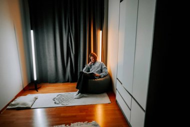 Genç bir kadın, yumuşak ışıklandırmalı bir odada bir koltukta otururken kitap okur.