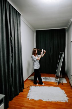 Aynanın önünde siyah perdeleri ve ahşap zemini olan bir kadın saç maşası kullanıyor.