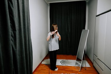 Sabah odasında aynanın önünde saç ütüsü kullanan bir kadın.