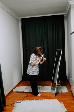 Kadın güne hazırlanırken aynanın önünde saç maşası kullanıyor.