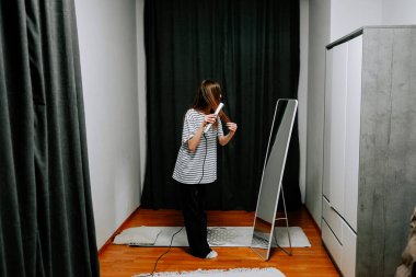 Kadınlar sade odada aynanın yanında dururken saçlarını demirle şekillendirirler.