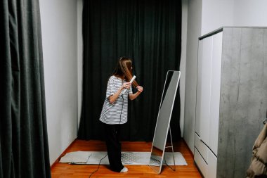 Güzel bir kadın saçını odasındaki aynanın önünde saç maşasıyla şekillendirir.