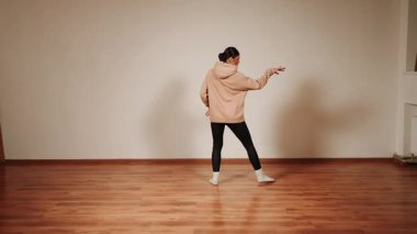 Kadın, basit bir dans salonunda dans eder. Neşe ve hareketlerini odaklanmış bir ifade tarzıyla gösterir.