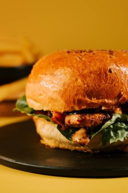 Parlak sarı bir arka planda siyah bir tabağa taze malzemeler konmuş sulu hamburger.