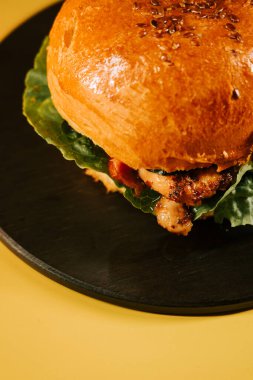 Siyah bir tabakta servis edilen sulu hamburger, sarı arka planda taze malzeme ve doku içeriyor.