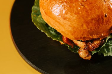 Sulu burger, parlak sarı bir arka planda taze malzemelerle siyah bir tabakta oturur.