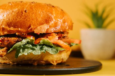 Salatalı ve domatesli sulu burger, bir bitkinin yanında sade sarı bir arka planda.