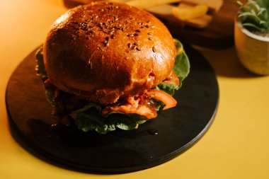 Siyah bir tabakta sulu burger, kıvırcık salata ve domates, parlak sarı bir arka planda.