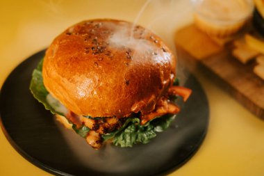 Sulu burger siyah bir tabakta dinleniyor, sarı bir zemin ve buhar yükseliyor.