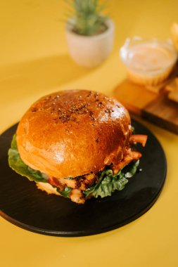 Sulu burger, sarı arka planda sebzeli siyah bir tabakta servis edilir.