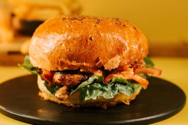 Sulu burger siyah bir tabakta oturur, sarı arka plan ve taze sebzelerle.