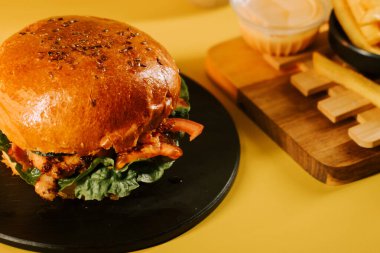 Siyah tabakta servis edilen marul ve domatesli sulu burger ve sarı arka planda patates kızartması.