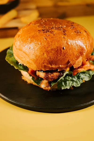 Sulu burger, parlak sarı bir arka planda, marul ve malzemelerle siyah bir tabakta oturur.