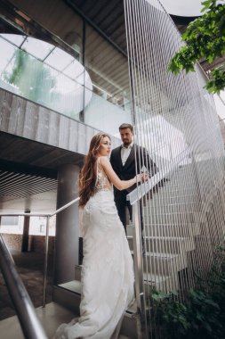 Resmi kıyafetli bir çift, öğleden sonra bir düğün töreninde modern bir mekanda merdivenlerden yukarı çıkıyor.