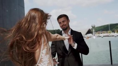 Yazın suya yakın dans eden çiftlerin arkasında tekneler ve yeşil tepeler var.