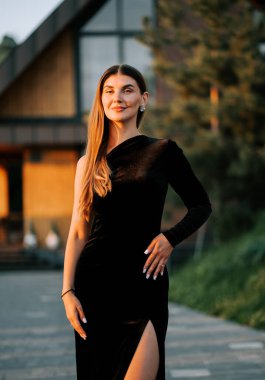 Model, gün batımında uzun siyah bir elbiseyle binanın önünde poz veriyor.
