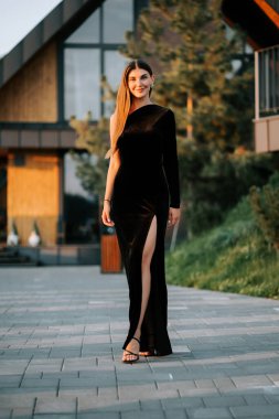 Modern bir binanın önünde duran zarif bir kadın. Uzun siyah elbisesi ve yan yarığı var.