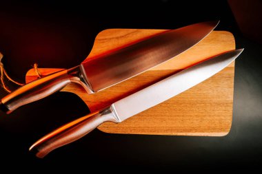 Akşam vakti karanlık bir mutfak ortamında ahşap bir kesme tahtasının üzerinde iki mutfak bıçağı.