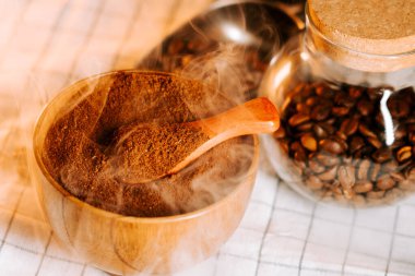 Tahta bir kasede öğütülmüş kahve buharı yükseliyor ve kahve çekirdekleri yakınlarda.