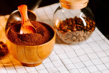 Tahta bir kasede öğütülmüş kahve kaşığı ve kahve çekirdekleri bir kavanozda masada.