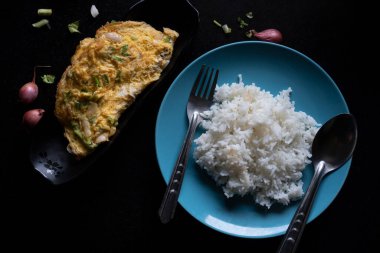 Yeşil soğan ve soğanla karıştırılmış omlet. Siyah bir tabakta ikiye katla. Mavi bir tabakta çatal ve kaşıkla temiz beyaz pirinçle servis yap. Arpacık soğanlı garnitür ve siyah fayanslı sebzeler..