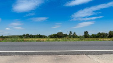 Tayland 'da asfalt bir yolun yatay ya da manzarası. Betonun önünde. Altın pirinç tarlaları hasat için hazır. Mavi gökyüzünün ve beyaz bulutların altındaki uzak yeşil orman bölgesi.