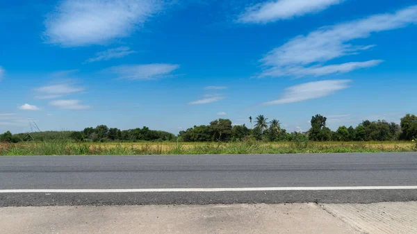 Tayland 'da asfalt bir yolun yatay ya da manzarası. Betonun önünde. Altın pirinç tarlaları hasat için hazır. Mavi gökyüzünün ve beyaz bulutların altındaki uzak yeşil orman bölgesi.