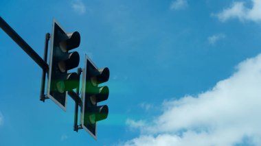 Yukarıya bak, çifte trafik ışığının yanlarını gösteriyor. Her iki tarafta da yeşil ışık görünüyor. Sinyaller geliyor. Mavi gökyüzünün altında ve gündüz beyaz bulutların altında.