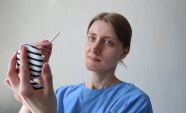 Genç bir kadın aynaya bakarken dudak parlatıcısı sürüyor. Doğal makyaj, evde güzellik rutini. Çizgili ayna ve dudak parlatıcısı tutan kadın. Cilt bakımı ve kendine bakım konsepti.