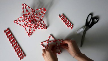 El yapımı. Kadın eli iplikle iğneyi tutuyor. Kokteyller için kağıt tüplerden Noel yıldızları yaratma sürecinde. Kırmızı beyaz çizgili pipetler. Makas, beyaz iplik. Beyaz arka plan. Fotoğraf.