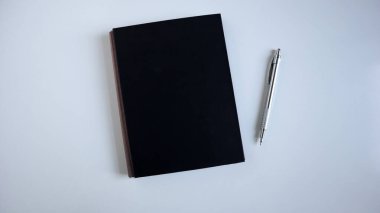 Minimalist düz taban, kapalı siyah bir defterin üst görünüm bileşimi ve temiz, aydınlık bir yüzey üzerindeki gümüş mekanik kalem. İş, eğitim, planlama, yaratıcılık, taklit sunumlar için ideal.