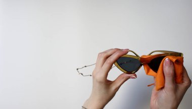 Beyaz arka planda turuncu mikrofiber bez ile güneş gözlüğü temizleme işlemi. Optik mağaza pazarlaması veya moda aksesuarları bakımı için minimalist stüdyo fotoğrafçılığı.