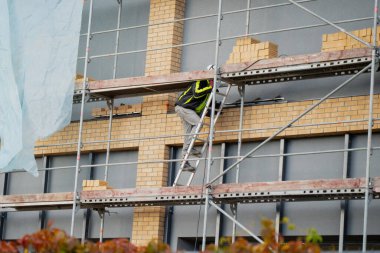 İnşaat işçisi tuğla şantiyeye tırmanıyor ön cephe kurulum sürecini, güvenlik ekipmanları kullanımını ve endüstriyel inşaat iş akışını gösteriyor