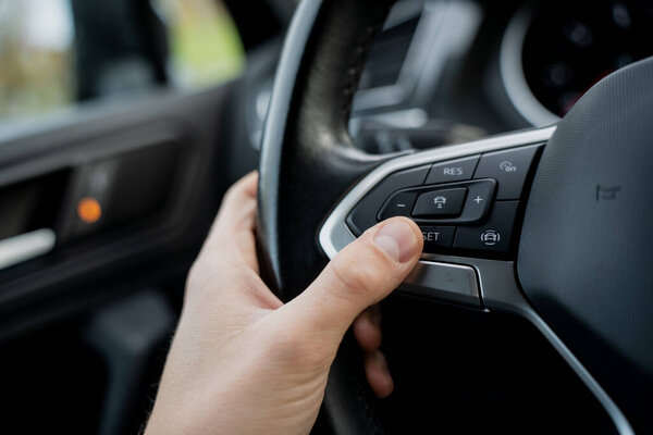 Driver hand pressing cruise control button on modern car steering wheel showing vehicle interior technology, driving comfort system and automotive control interface detail