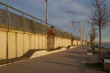 Riga 'daki boş nehir kenarı gezinti alanı, sıcak akşam güneşi altında banklar, ağaçlar ve sel duvarı huzurlu şehir atmosferini, kentsel tasarımı ve deniz kenarındaki manzaralı yaya geçidini gösteriyor.