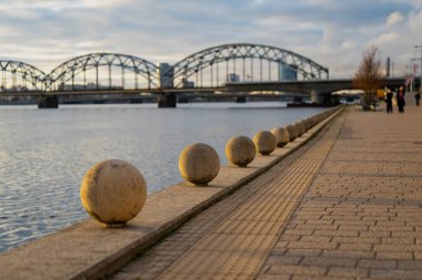 Riga 'da granit küre bariyerleri ve arka planda Daugava demiryolu köprüsü olan nehir kenarı geçidi manzaralı, sakin gün ışığında kentsel peyzaj tasarımı ve şehir mimarisini gösteriyor
