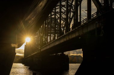 Riga demiryolu köprüsünün gün batımında altyapı tasarımı, çelik iskeleti ve Letonya 'nın endüstriyel mühendislik ve mimari bileşimini gösteren suyun ışık yansımalarını gösteren alt köprü görüntüsü