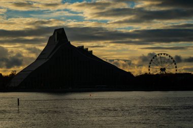 Gün batımında Riga şehir manzarası silueti, Daugava Nehri 'nin yanındaki Ulusal Letonya Kütüphanesi ve Dönme Dolap' ı kasvetli gökyüzü ve şehir kültürünü ve mimari kimliğini gösteren sıcak bir ışıkla gösteriyor.